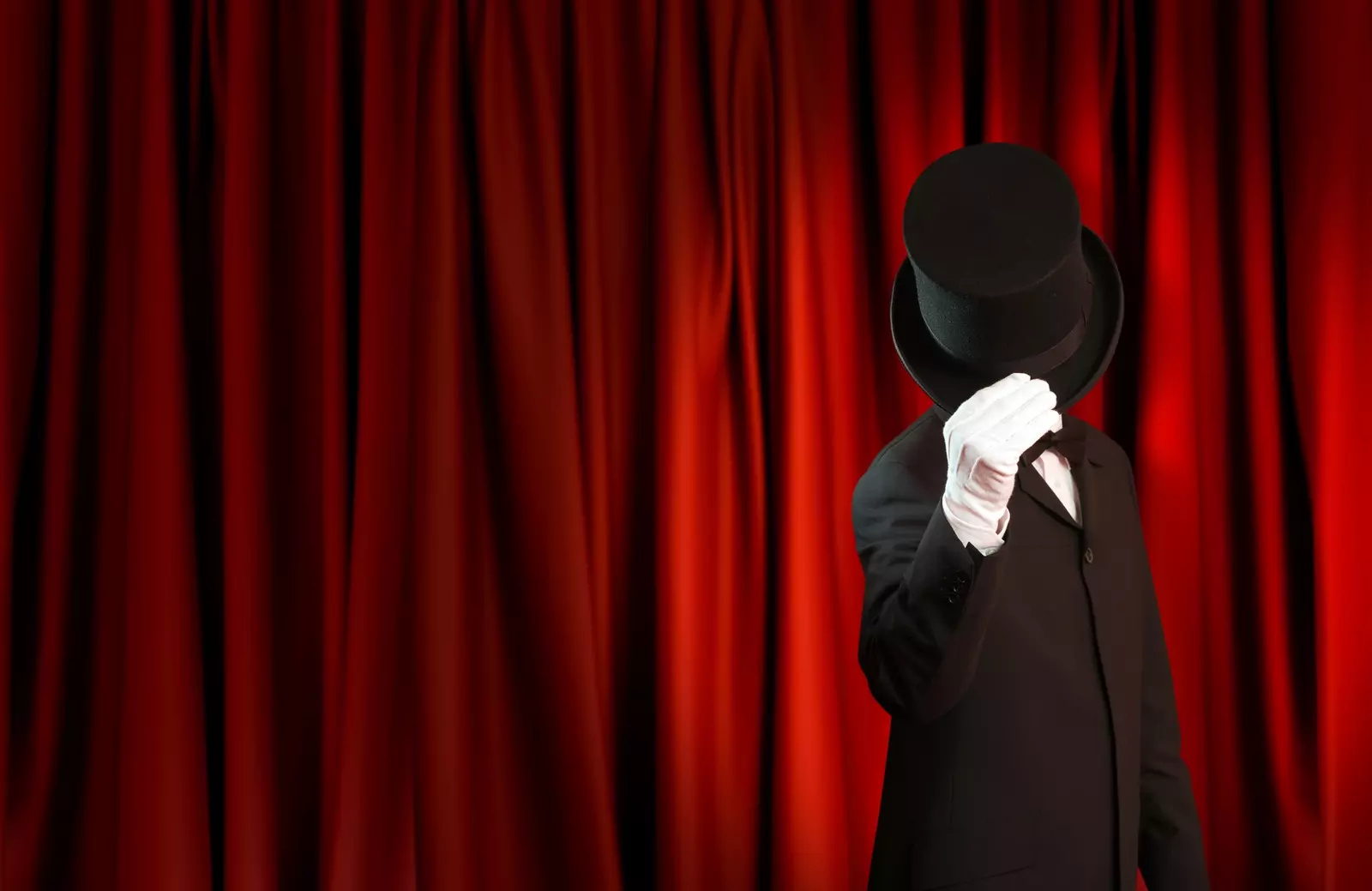 A tuxedoed magician is standing in front of a red curtain. His hat is tipped over his face.