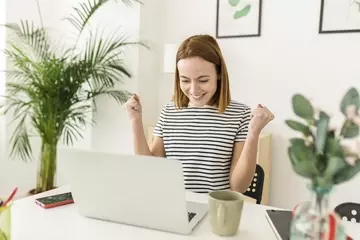 Cheerful young woman celebrating her achievement while reading good news on laptop from home office - Smiling female expressing excitement working at home