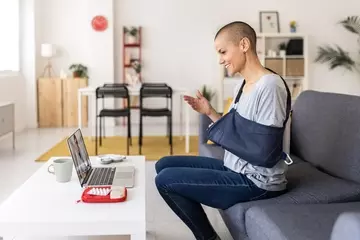 A woman sitting on a couch with her arm in a sling. She is looking at her mobile phone and has a laptop on the coffee table in front of her. 