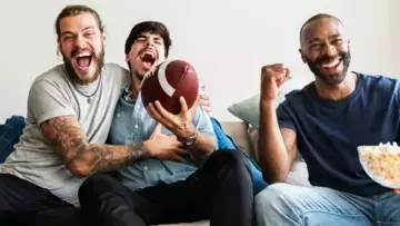 Three men cheering and celebrating enthusiastically on a couch. The man in the center is holding football.