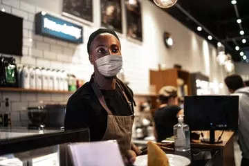 Hourly employee working at cafe standing masked behind counter